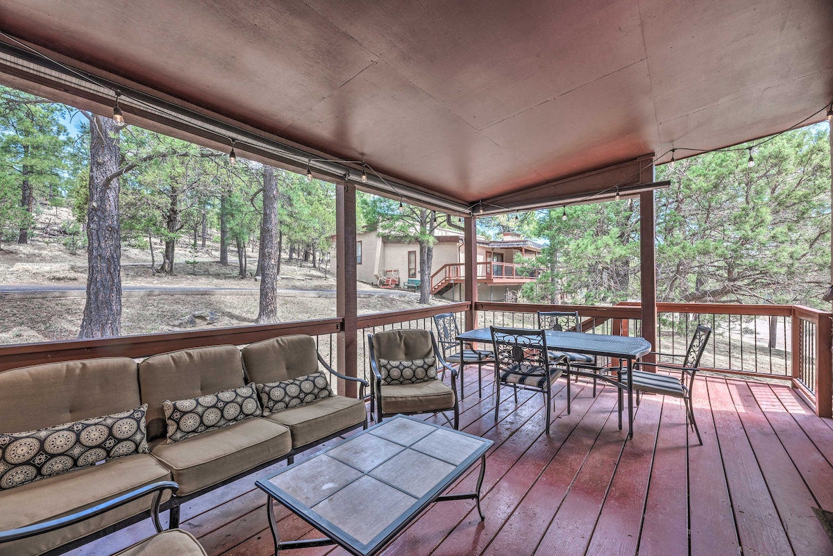 A spacious covered porch is furnished with multiple seating options, including a sectional sofa and dining table. The surrounding trees provide shade, enhancing the natural setting. The flooring is wooden, and large windows allow for ample light and views of the outdoors.
