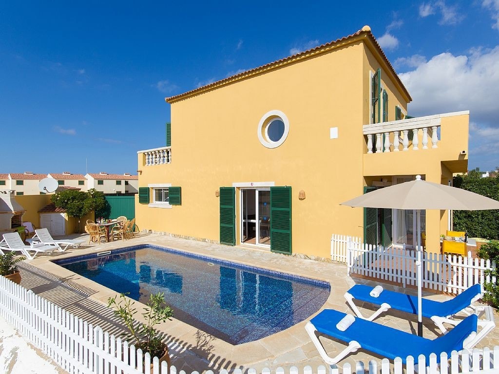 A bright yellow villa is showcased, featuring large green shutters and round windows. A private swimming pool is surrounded by a sun terrace with loungers and a parasol. The property is enclosed by a low white fence, providing a welcoming outdoor space.