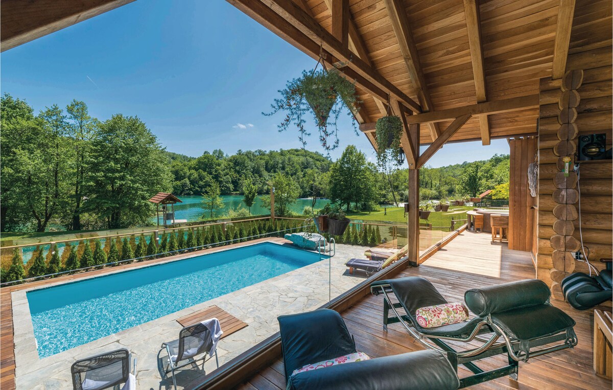 An outdoor area features a swimming pool framed by a wooden deck, with lounge chairs positioned for relaxation. Lush greenery surrounds the space, providing a scenic view of the river and landscape beyond. Light streams in through expansive glass doors, creating an inviting atmosphere.