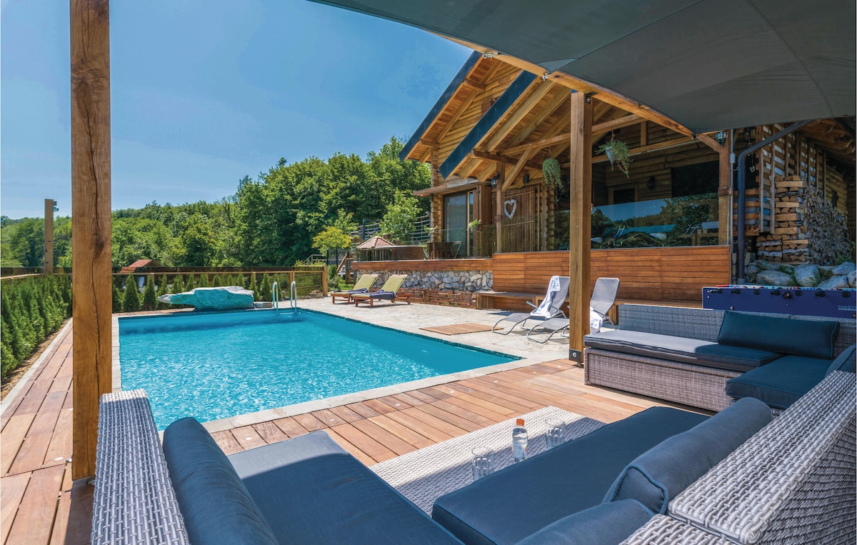 The outdoor area features a rectangular swimming pool surrounded by a spacious wooden deck. Comfortable loungers are arranged nearby under a shaded area, while the wooden structure of the holiday home is visible in the background, set against lush greenery.