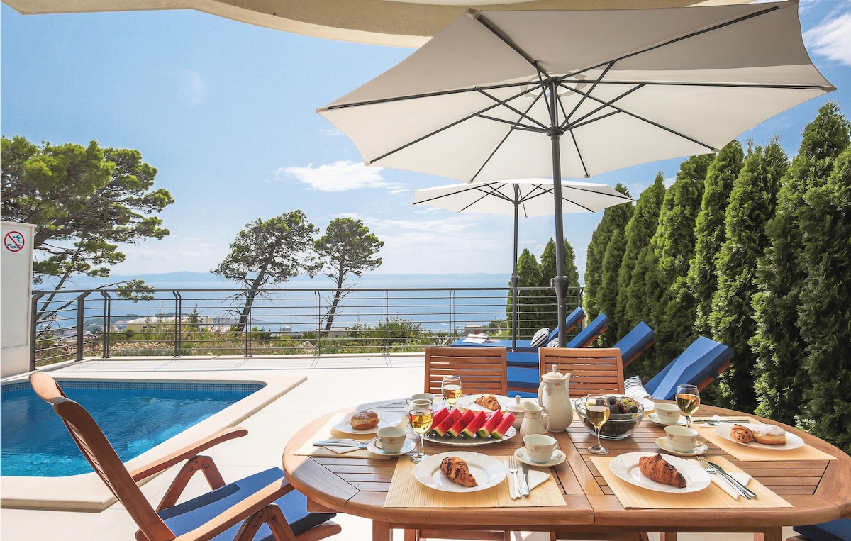 An outdoor dining area is displayed, featuring a wooden table set with breakfast items, including croissants and fruit. Sun loungers and umbrellas surround a private pool, with a view of the sea and trees in the background. A peaceful setting invites relaxation.