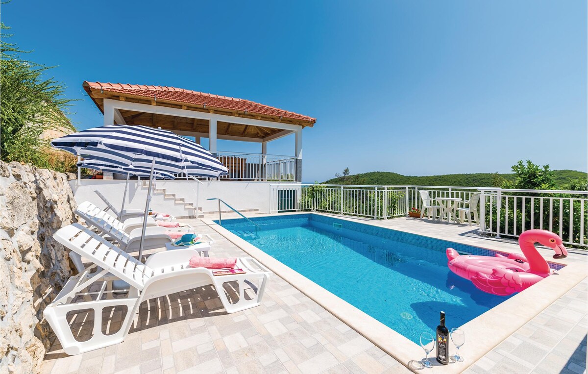 An outdoor pool area is framed by a stone wall, featuring lounge chairs arranged beside the inviting turquoise water. A striped umbrella provides shade, and a pink inflatable flamingo floats nearby. A covered terrace can be seen in the background, offering additional seating.