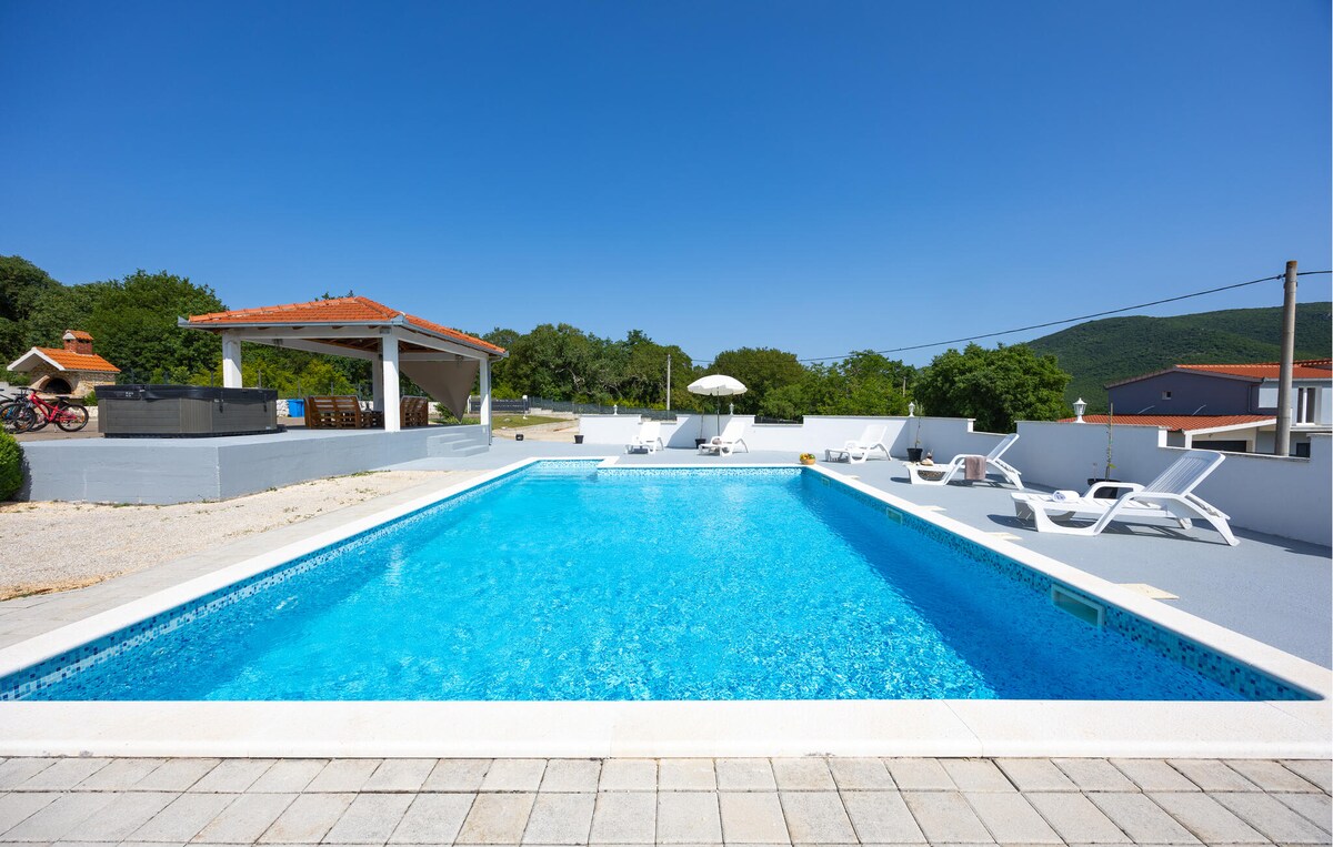 An outdoor private pool is visible, surrounded by a sun terrace. Several lounge chairs are placed around the pool. A shaded area is provided by an umbrella, and a structure for dining or relaxation is seen in the background, framed by green trees.