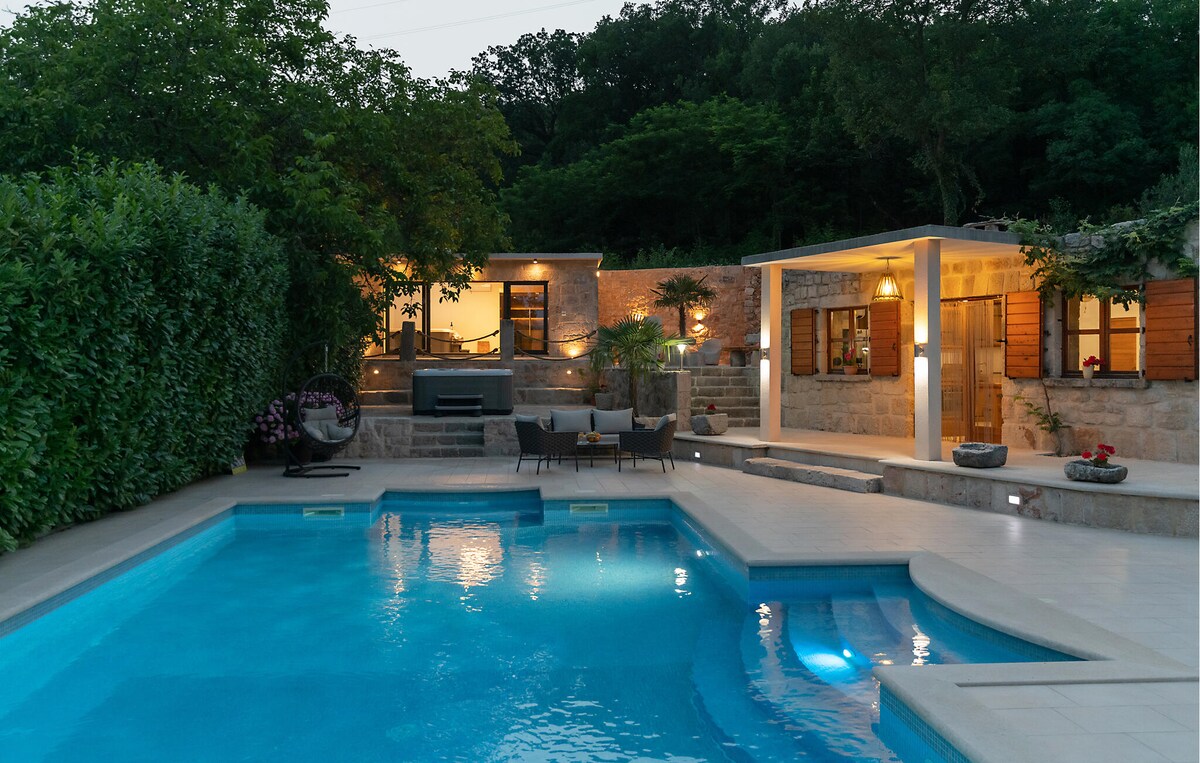 The inviting outdoor area features a sparkling pool surrounded by lush greenery and stylish poolside seating. The stone structure in the background includes a covered patio area with warm lighting, creating a relaxing ambiance for evenings by the pool.