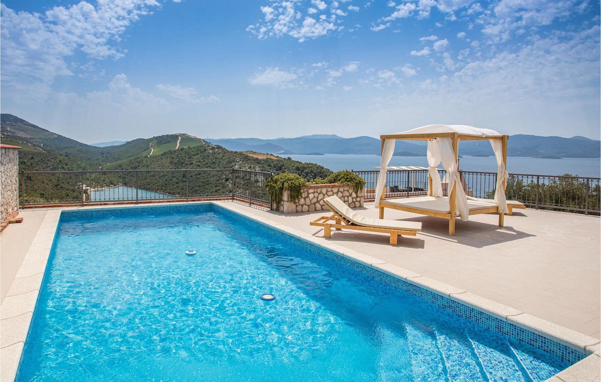 An outdoor pool is surrounded by a spacious patio featuring a sun lounger and a shaded cabana. Panoramic views of the sea and green hills extend beyond the pool, under a clear blue sky dotted with wispy clouds.