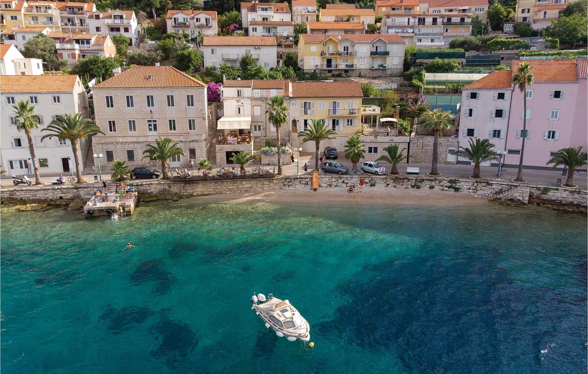 A coastal view features a sandy beach lined with palm trees and quaint buildings in pastel colors. A boat floats gently in the clear, turquoise water, while a small pier extends into the sea. Lush greenery and blooming flowers add to the tranquil seaside scenery.