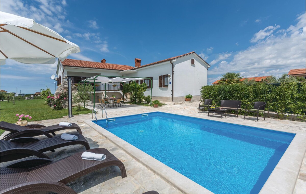 A private outdoor pool, surrounded by a spacious stone patio, is complemented by several sun loungers. A shaded area with tables and chairs is positioned nearby, with the house and gardens visible in the background under a clear blue sky.