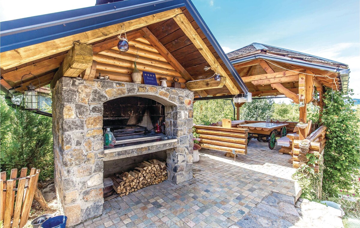 An outdoor gazebo is depicted, featuring a stone barbecue area with wood stacked nearby. Rustic wooden benches and tables provide ample seating, surrounded by lush greenery. A covered section extends the space, offering shelter while enjoying nature.