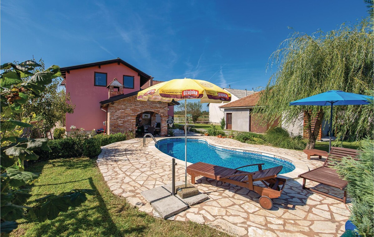 A private outdoor pool is framed by a stone patio and surrounded by well-maintained grass. Colorful umbrellas provide shade over lounging chairs. A pink house with multiple windows stands in the background, complementing the clear blue sky.