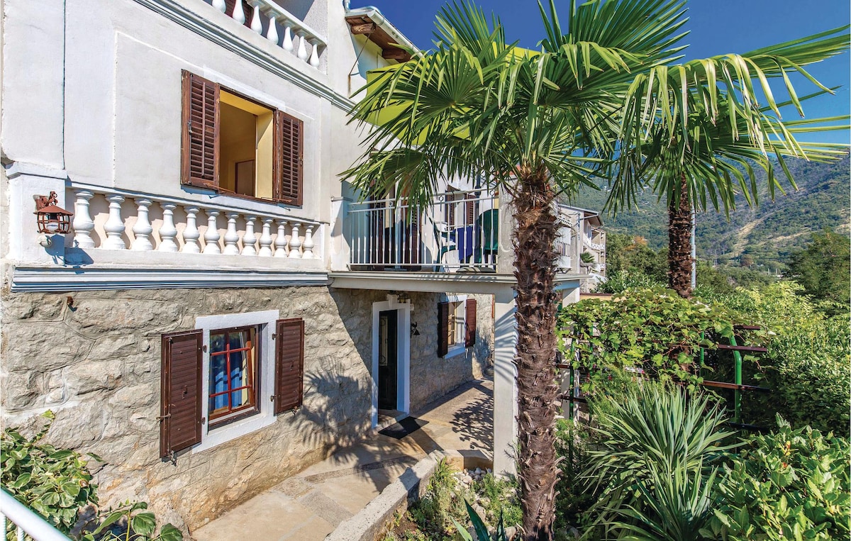 The exterior of the two-story holiday house is showcased, featuring stone walls and decorative shutters. Lush palms and greenery surround the entrance, while a balcony extends from the upper floor. Bright blue window panes add color to the inviting facade under a clear blue sky.