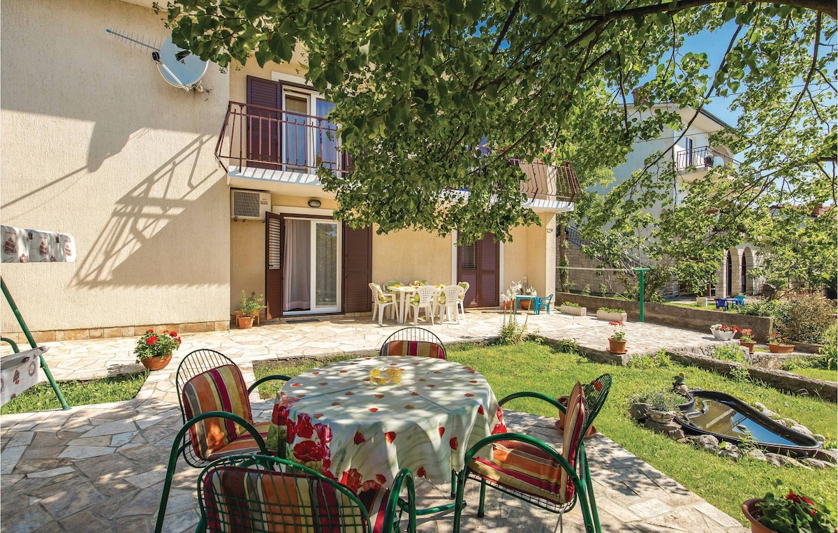 A spacious terrace is presented, surrounded by greenery, featuring a round table covered with a patterned cloth. Colorful chairs are arranged around the table, with potted plants enhancing the landscape. The background showcases the beige façade of the house and sliding glass doors leading indoors.