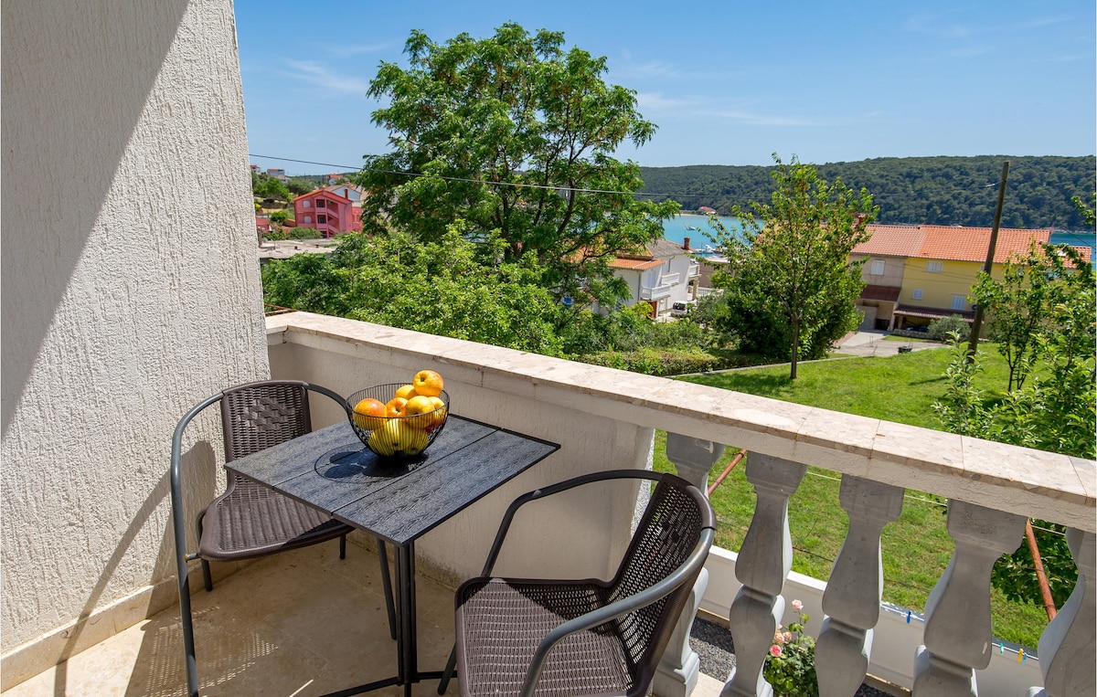 A balcony is visible, furnished with two black chairs and a glass table, holding a bowl of oranges. Overlooking the countryside, the view includes greenery, a body of water, and distant structures, contributing to a serene outdoor setting.