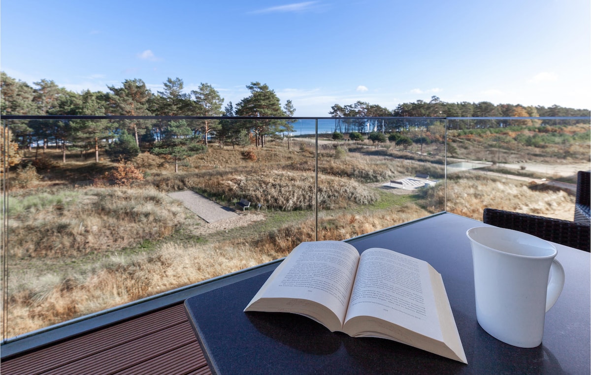 A spacious balcony features a table with an open book and a cup, overlooking a natural landscape with grassy areas and trees. The calm blue waters of the Baltic Sea can be seen in the distance, providing a serene backdrop.