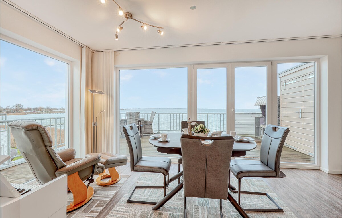 A dining area is featured with a round table set for four, surrounded by sleek chairs. Large windows provide panoramic views of the water and surrounding nature, while a cozy armchair is positioned nearby. Bright, natural light illuminates the space.