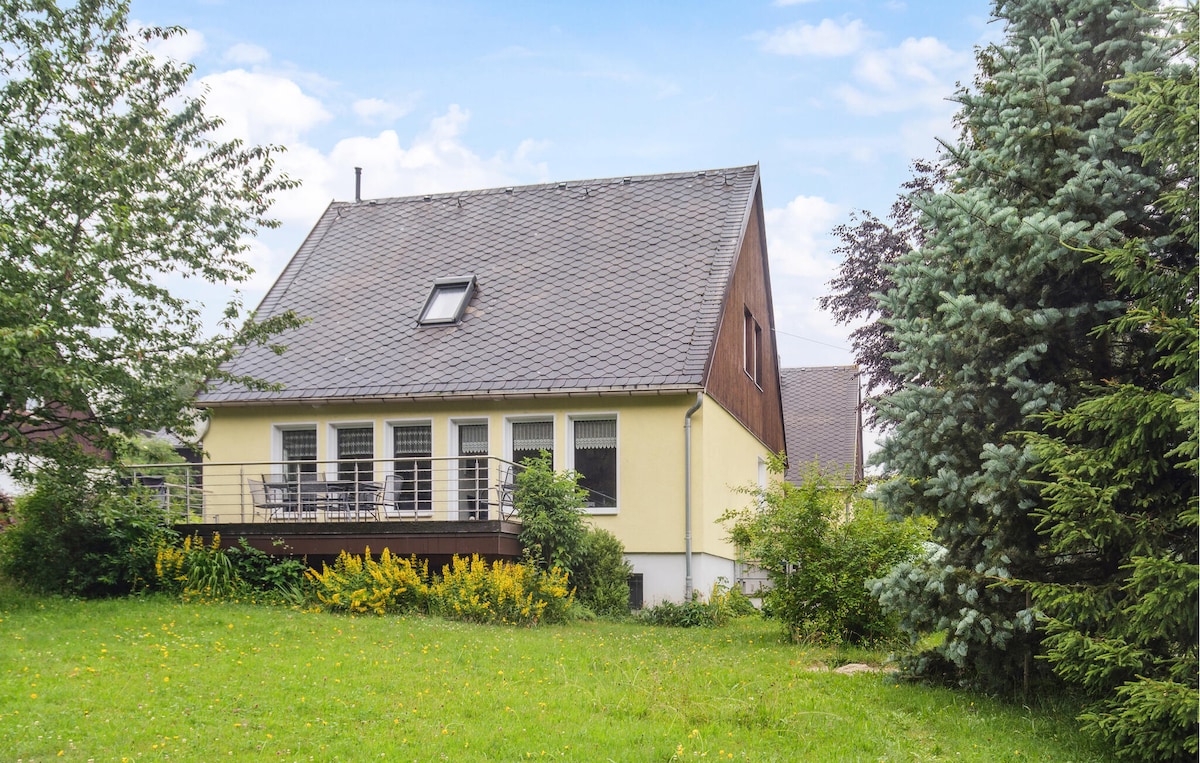 A two-story vacation home is set amidst a green lawn, surrounded by trees and flowering shrubs. The exterior features a balcony with seating, offering a view of the garden. The roof is sloped, and large windows provide natural light to the home.