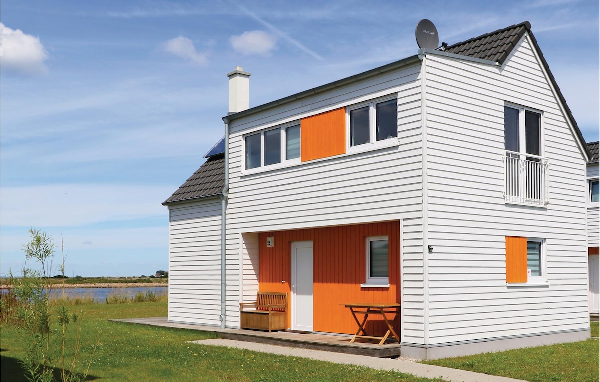 A two-story holiday home features a bright exterior with white siding and orange accents. The structure includes a small balcony on the upper level and a welcoming outdoor space with a bench and table. Lush green grass surrounds the house, creating an inviting entry.