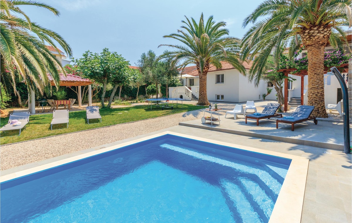 A private outdoor pool surrounded by palm trees is displayed. Lounge chairs are neatly arranged on the deck, while a grassy area with trees and a gazebo can be seen in the background. The overall setting provides an open and inviting outdoor space.
