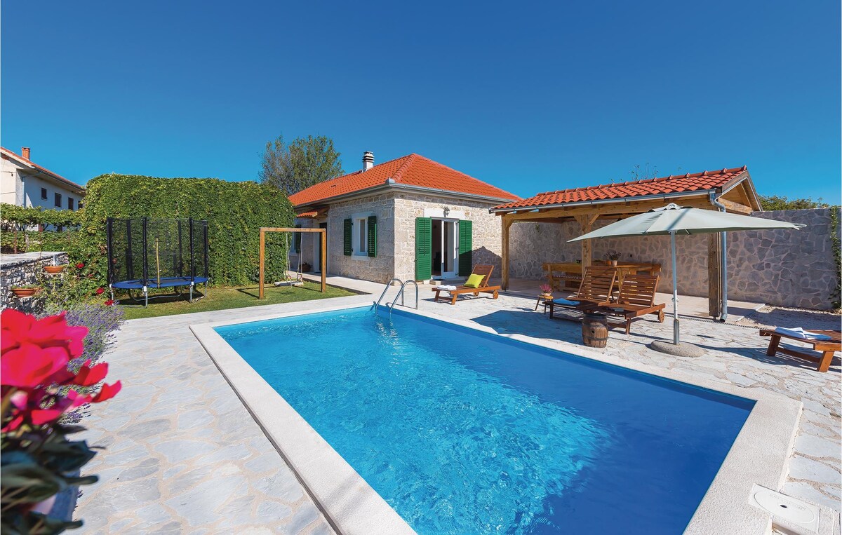 A stone house is featured beside a private outdoor pool, surrounded by greenery. Sun loungers and a shaded seating area are positioned near the pool. A trampoline and wooden play structure are visible in the background, creating a welcoming outdoor space.