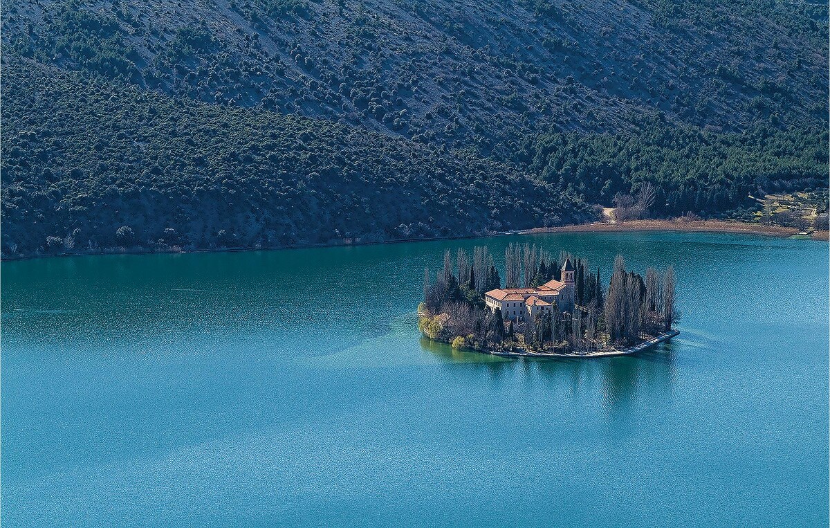 An aerial view captures a serene island surrounded by calm blue waters. A cluster of trees and a traditional stone building are visible, offering a sense of tranquility. The distant hills are adorned with lush greenery, enhancing the peaceful setting.
