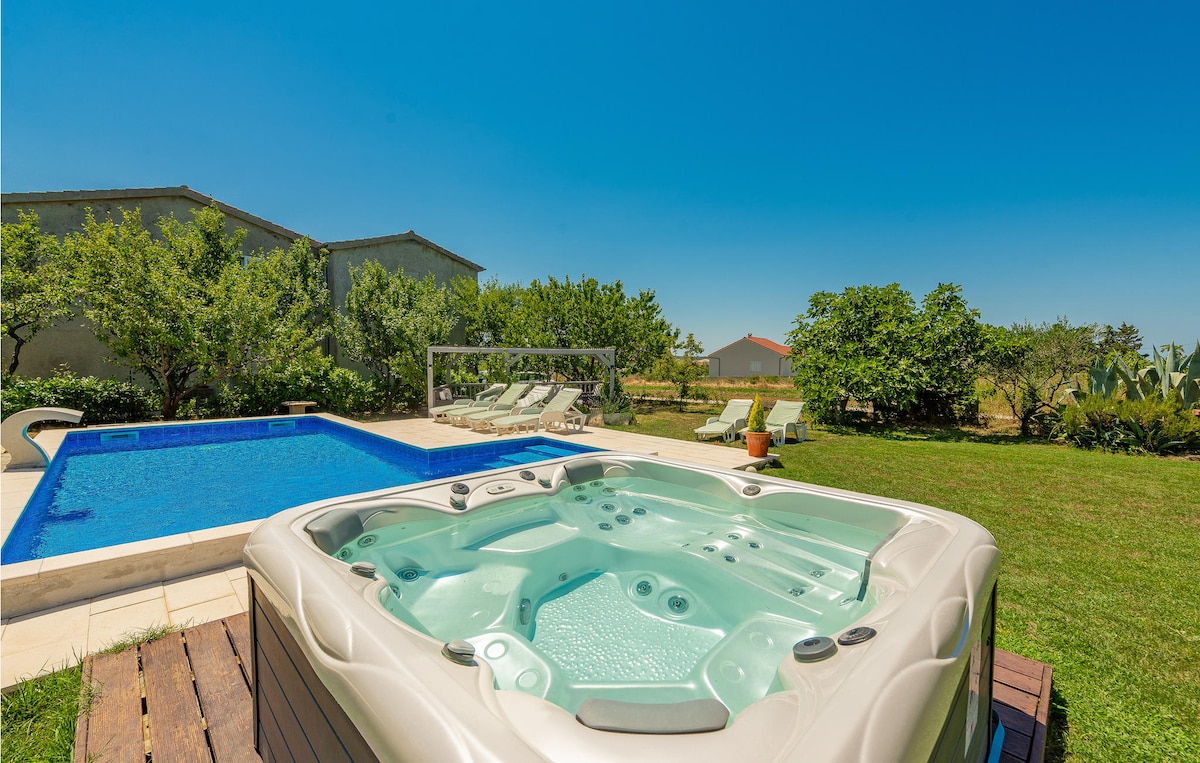 A private outdoor area is featured, showcasing a hot tub adjacent to a swimming pool. Lounge chairs are arranged around the pool, surrounded by lush greenery and trees. A clear blue sky complements the serene atmosphere of this space.