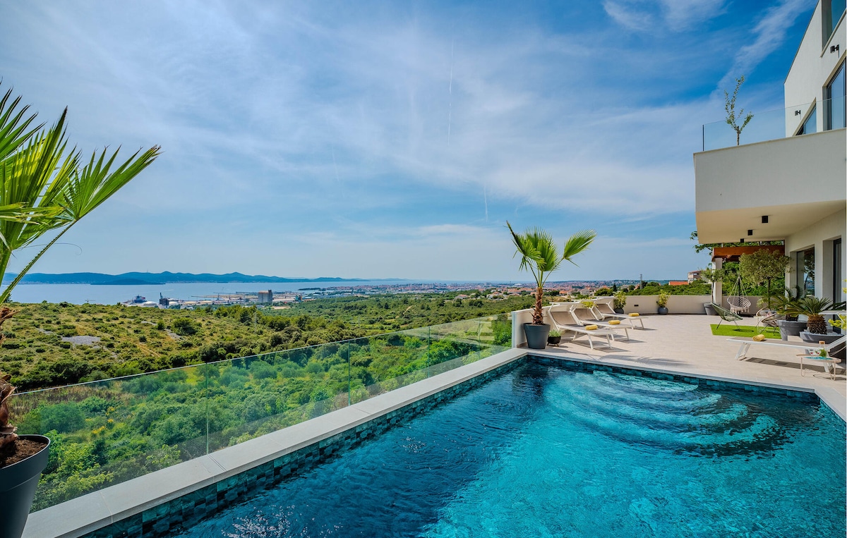 An outdoor private pool is showcased, bordered by a smooth stone patio. Sun loungers and tropical plants are visible in the background. A panoramic view of the sea and the Zadar archipelago can be observed, with clear skies enhancing the scenery.