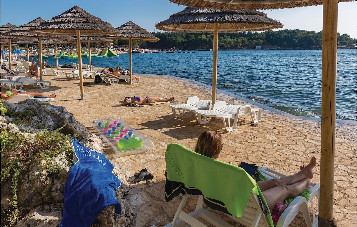 A sunlit beach area features several lounge chairs under thatched umbrellas, providing shaded spots for relaxation. A calm sea reflects the bright sky, and a colorful inflatable float rests on the sandy edge. The atmosphere suggests a perfect place for enjoying nature by the water.