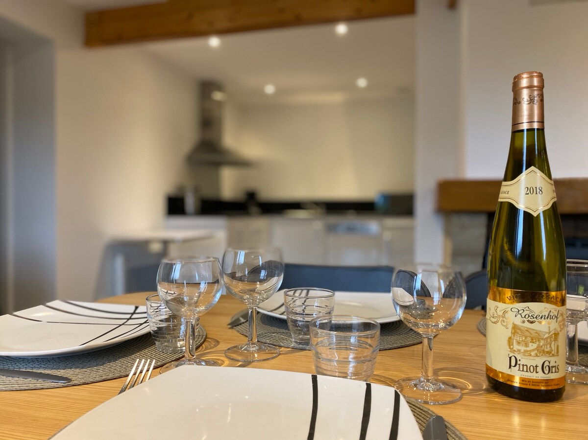 A dining table is set with white plates featuring black striped designs, accompanied by glassware, including wine glasses and tumblers. A bottle of Pinot Gris stands prominently on the table. The background reveals a modern kitchen with sleek cabinetry and appliances.
