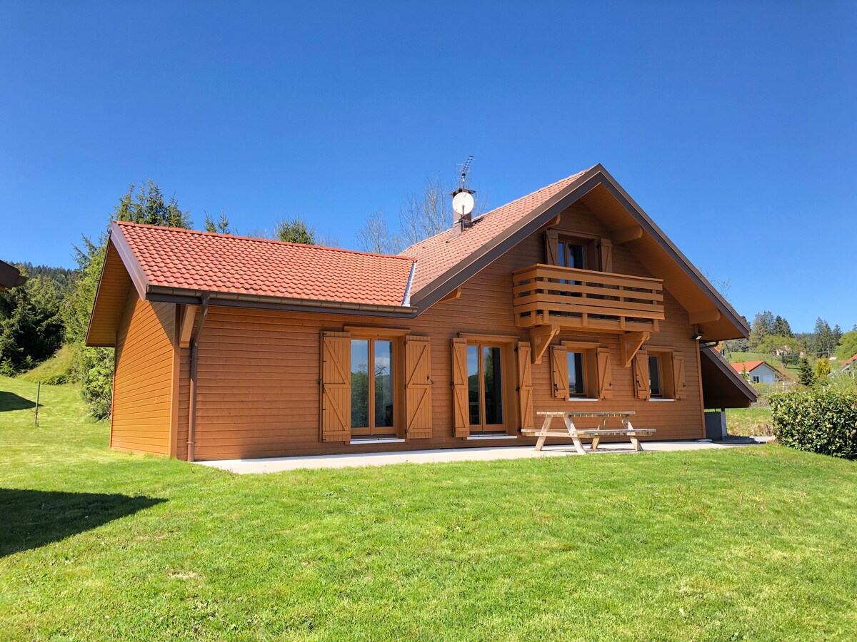 The exterior of the chalet is depicted, featuring a warm wooden facade with a tiled roof. A spacious patio area is visible, and a picnic table is situated on the lush green lawn. Surrounding trees and a clear blue sky enhance the serene outdoor setting.