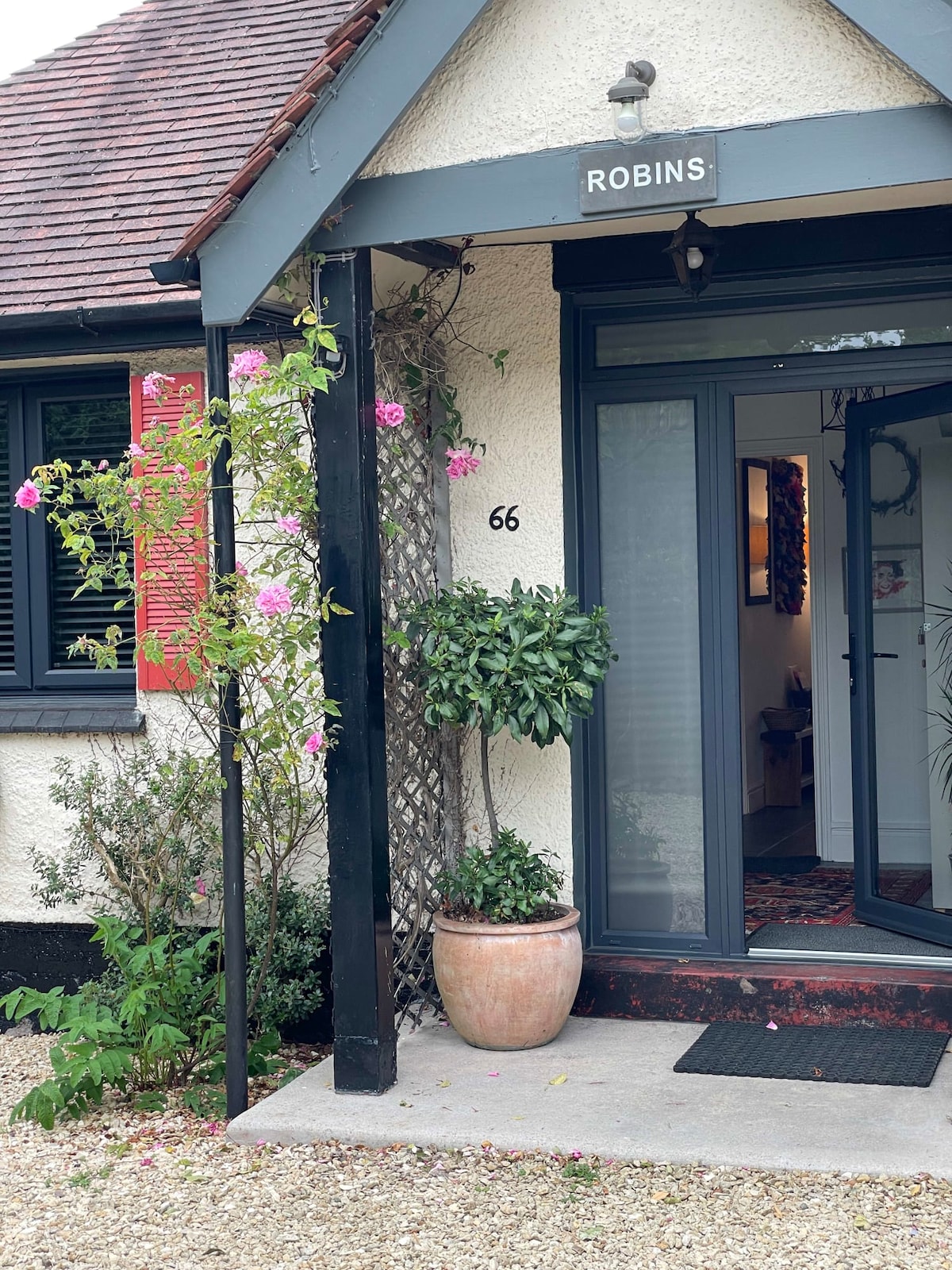 The entrance to the cottage features a welcoming doorway with the name 'Robins' displayed above. A potted plant and blooming vines adorn the entrance, complemented by a stone pathway and decorative gravel.