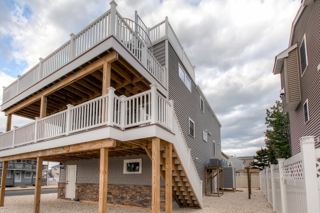 The exterior of the vacation rental is showcased, featuring a multi-level structure with a wooden deck supported by pillars. A staircase leads to the upper level, and the building is surrounded by a gravel yard and a white fence. Cloudy skies provide a soft backdrop.