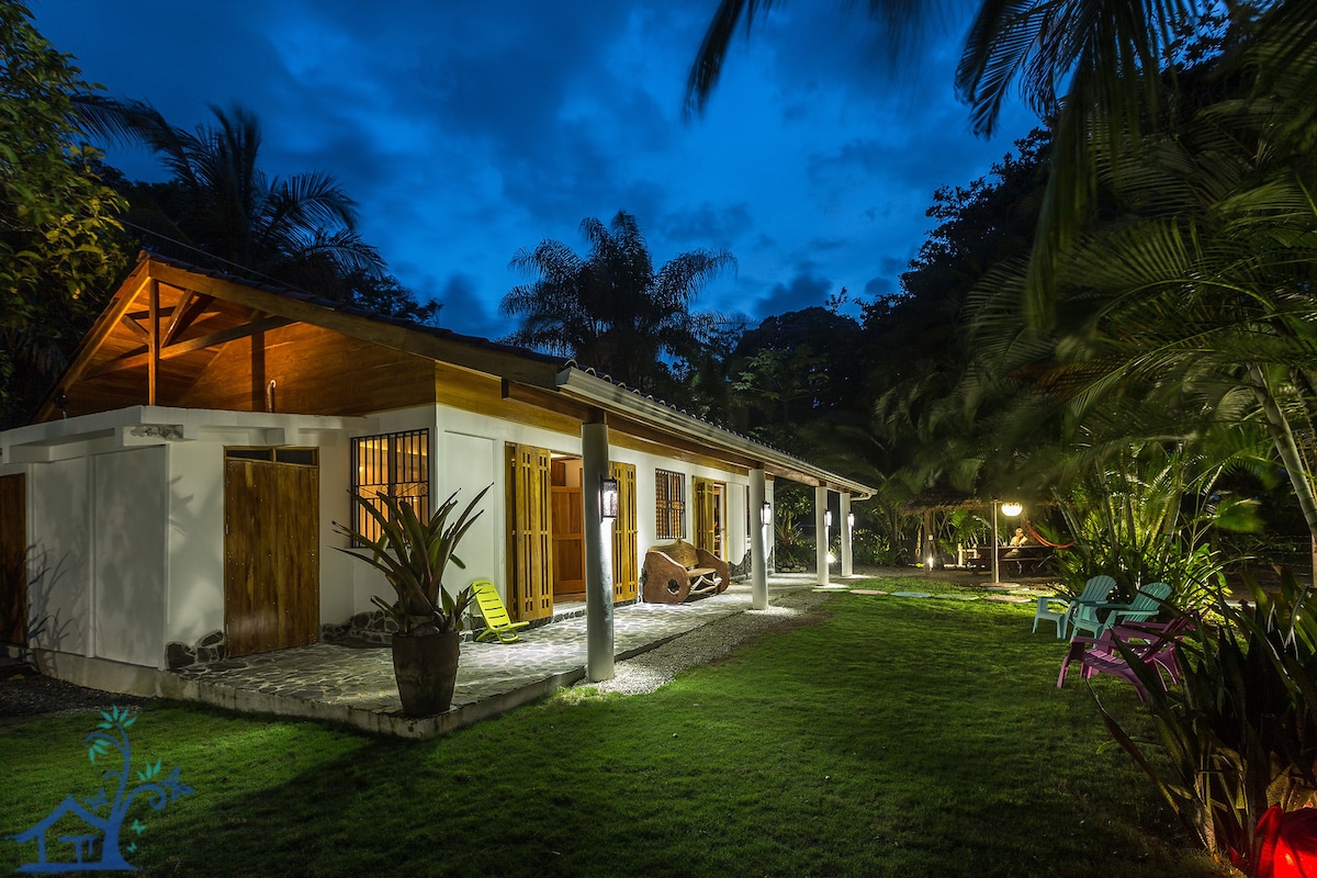 The bungalow is set amidst lush tropical landscaping, illuminated softly by exterior lighting at dusk. The structure features a spacious veranda with multiple seating options, green lawn, and a mix of tropical plants enhancing the outdoor space.