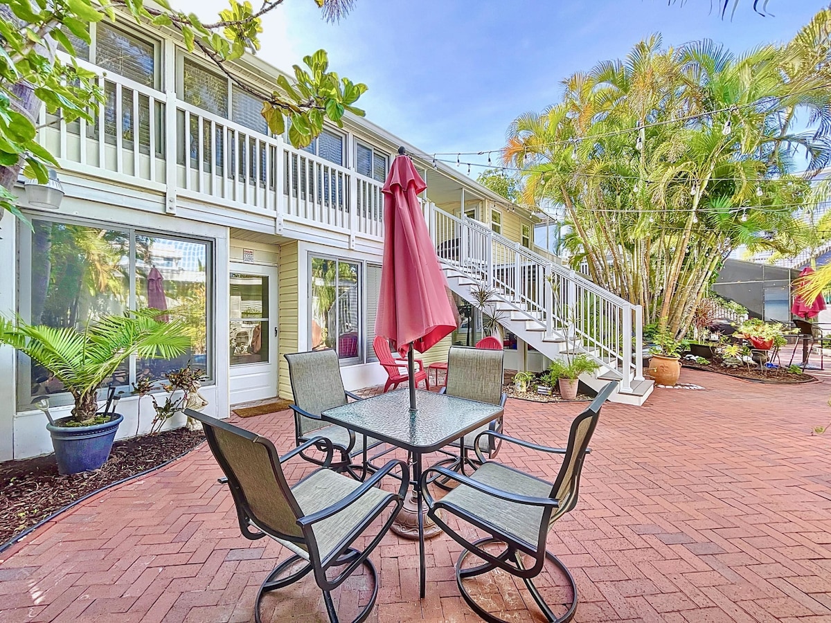 A private courtyard is highlighted by a dining set featuring a glass table and four chairs under a red umbrella. Lush greenery surrounds the space, and a flight of stairs leads up to the entrance of the building, with large windows providing natural light.