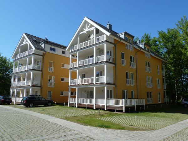 A bright, multi-story building is presented, featuring balconies with white railings on each level. The exterior is painted in a vibrant yellow, surrounded by lush greenery. A gravel parking area is visible in the foreground, accommodating several parked cars.