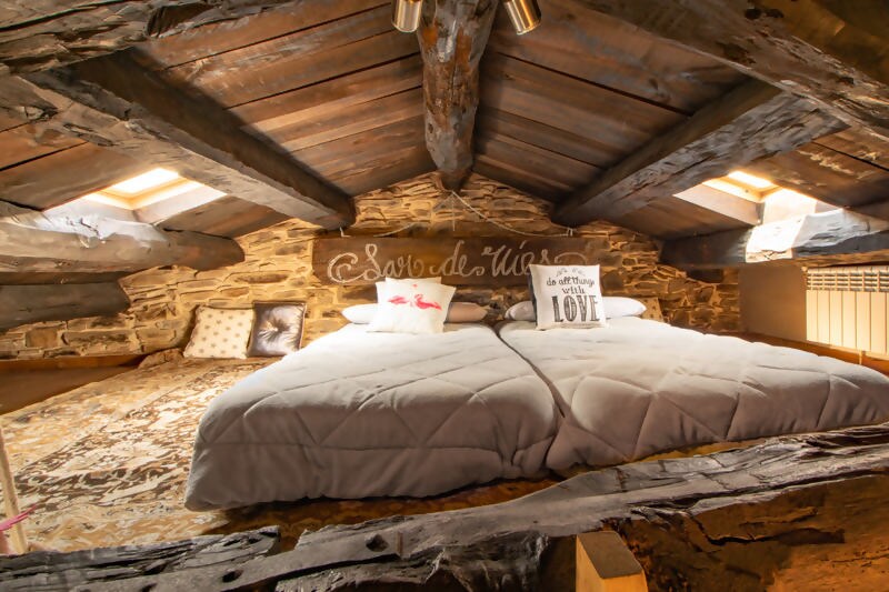 An inviting loft area showcases wooden beams and stone walls, featuring two spacious beds dressed in gray comforters. Skylights above provide natural light, illuminating the cozy sleeping space decorated with pillows and a charming 'Love' sign.