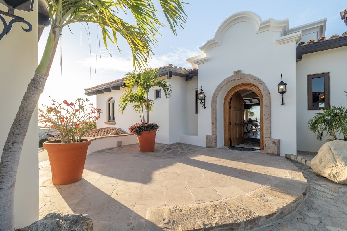 An inviting entrance of the villa is framed by lush plants and decorative pots. The stone pathway leads to a grand arched doorway, welcoming guests into the home. Natural light casts gentle shadows on the textured stone floor.