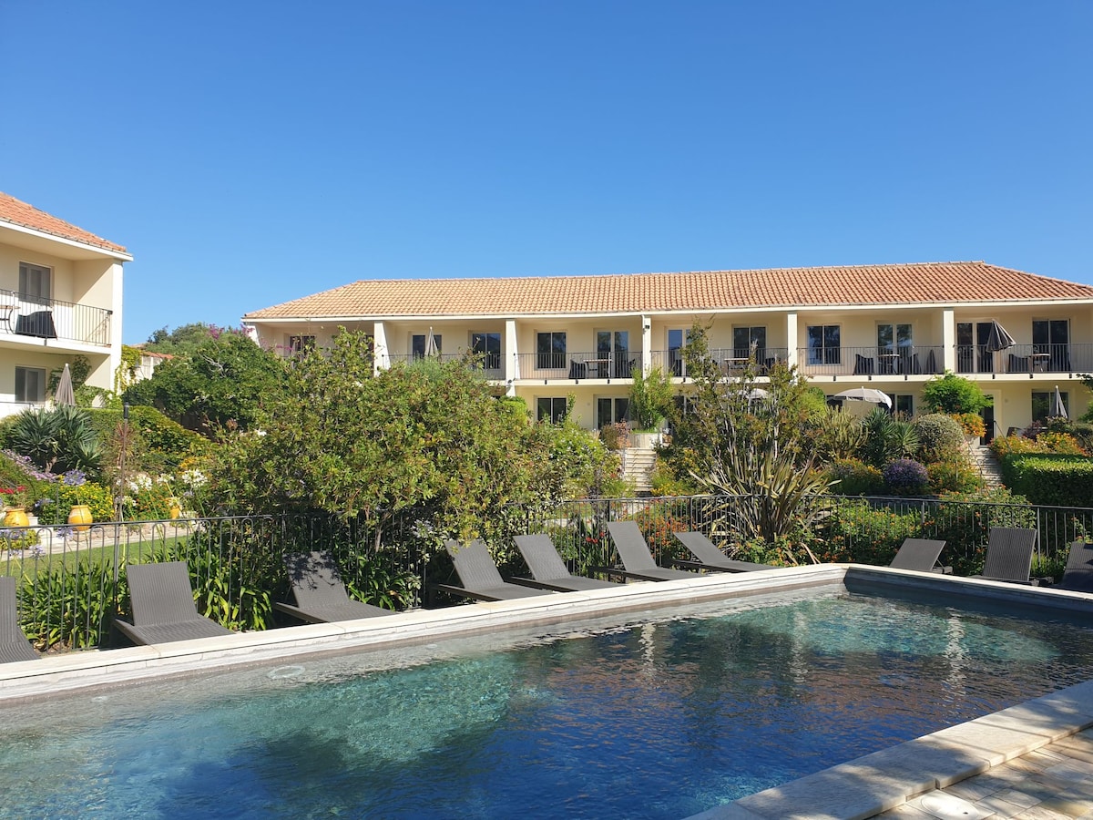A peaceful resort scene showcases a clear swimming pool surrounded by sun loungers. Lush greenery and flowering plants are seen throughout the garden, with two-story buildings visible in the background under a clear blue sky.