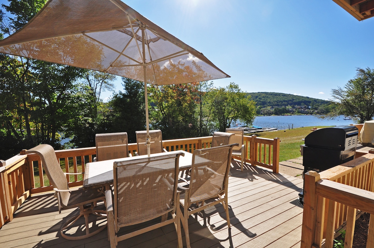 A spacious outdoor deck is shown featuring a large table surrounded by six chairs, with a canvas umbrella providing shade. A gas grill is positioned in the corner, and a scenic view of the lake is visible in the background, framed by greenery.