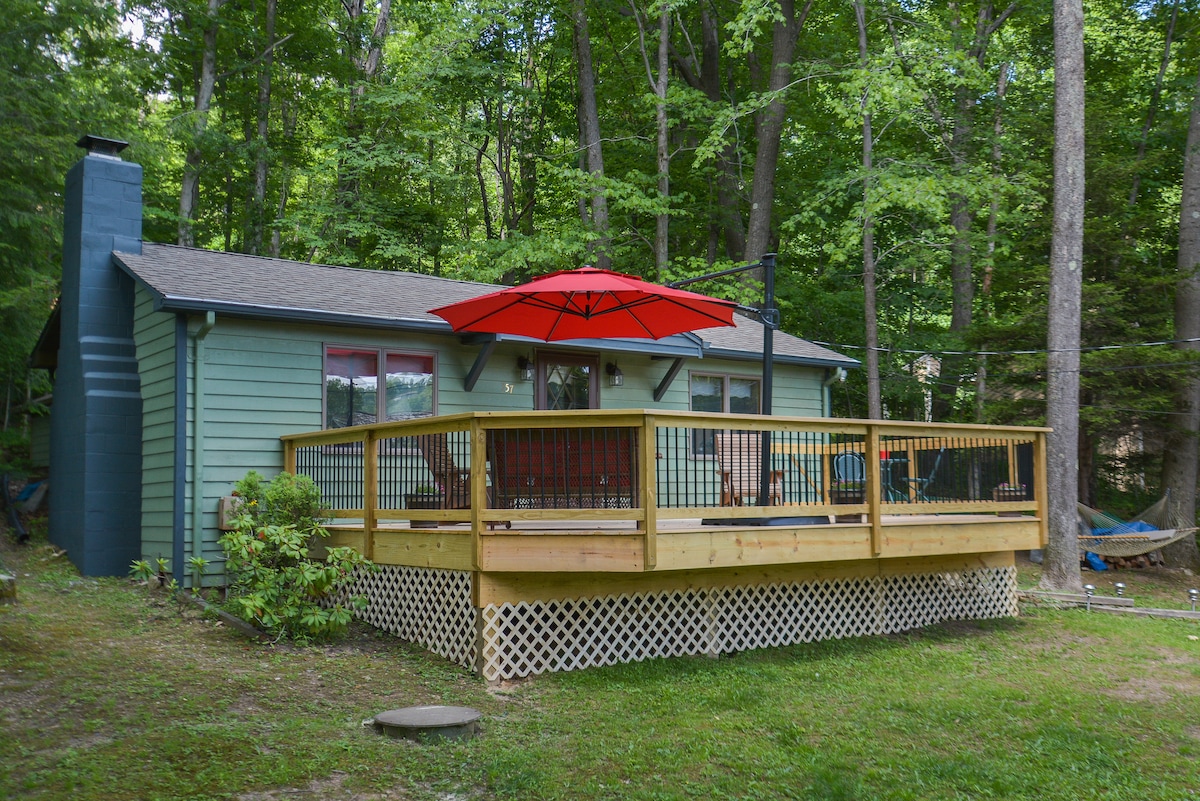 A cozy cottage is surrounded by lush greenery, featuring a spacious wooden deck. A bright red umbrella provides shade over outdoor seating, with large windows inviting natural light into the interior. A hammock can be seen nearby, enhancing the serene outdoor environment.
