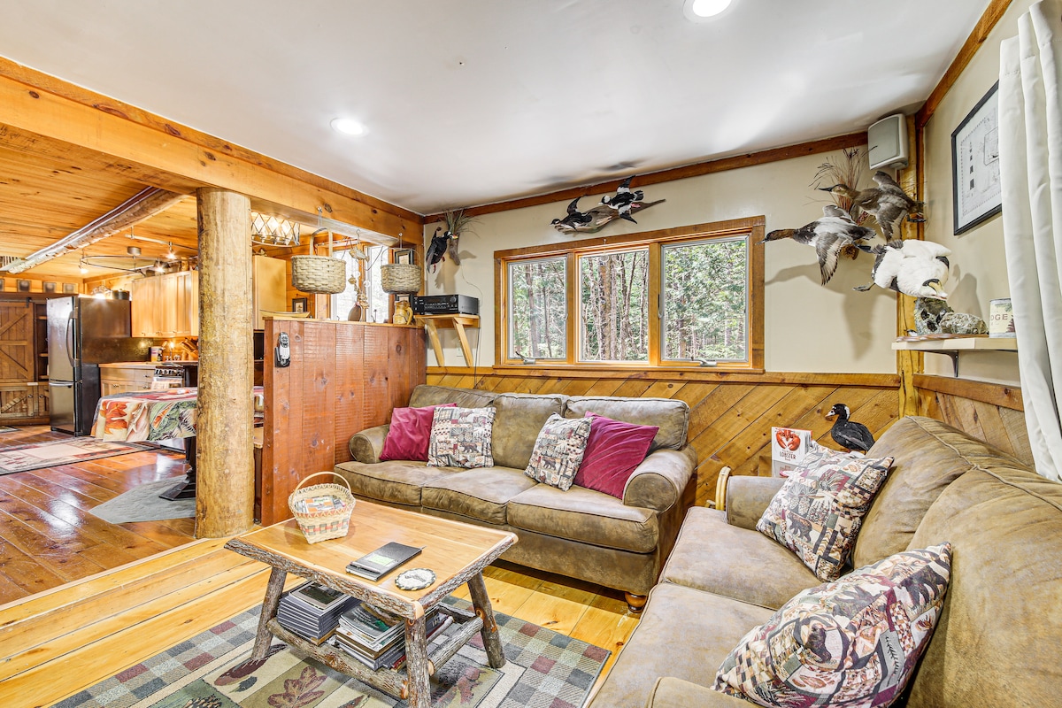 A comfortable living area features two plush sofas adorned with decorative pillows. A wooden coffee table is positioned between them, holding magazines. Natural light filters through windows, offering views of the surrounding forest. Rustic wood accents and artful decorations enhance the cabin's ambiance.