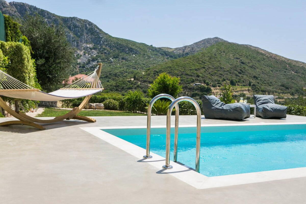 A refreshing swimming pool is framed by a smooth, light-colored patio. Two comfortable lounge chairs are positioned nearby, along with a hammock suspended between two supports. Lush greenery and rolling hills are visible in the background, depicting a serene natural setting.