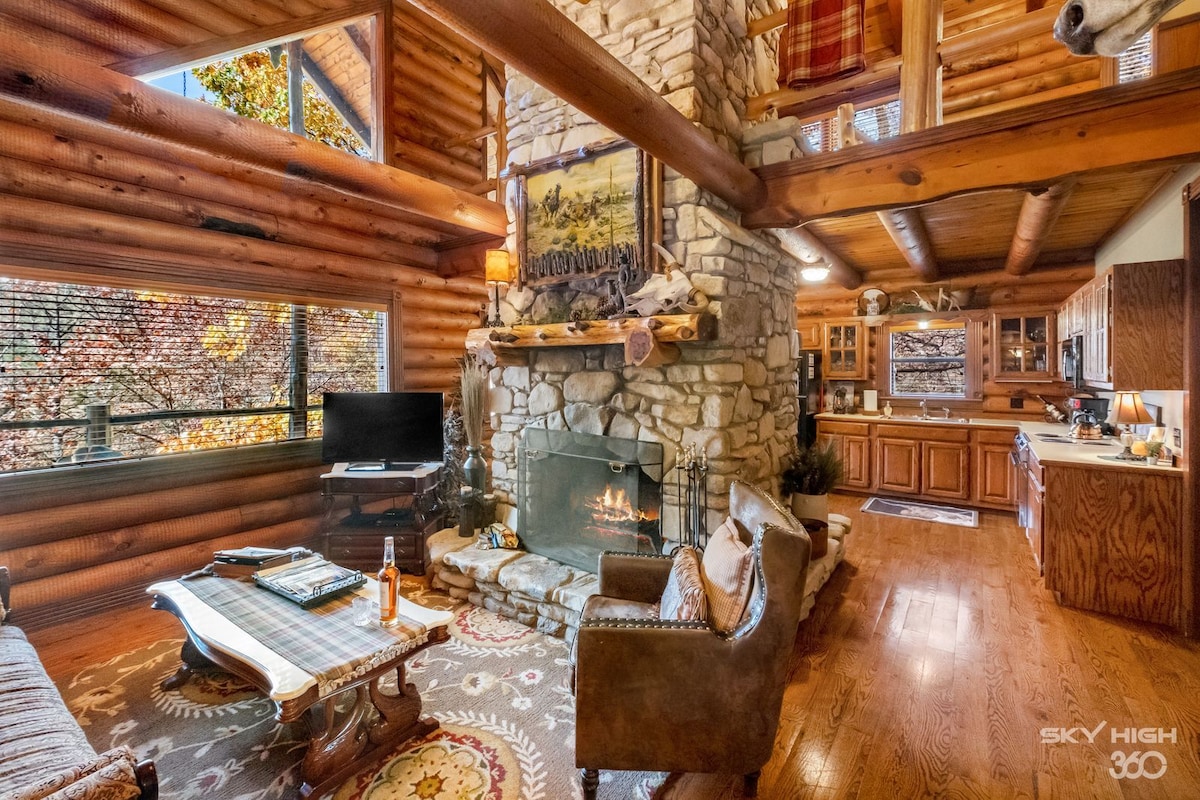 A spacious living area features a large stone fireplace as a focal point, with comfortable seating arranged nearby. Large windows allow natural light to fill the space, offering views of the surrounding landscape. The kitchen area is partially visible, showcasing wooden cabinetry.