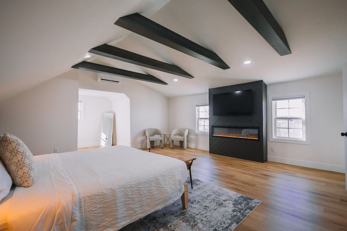 A spacious bedroom is illuminated by natural light, featuring a large bed with crisp linens. Dark wooden beams accentuate the ceiling, while a mounted television and an electric fireplace offer modern comfort. Two chairs and a mirror complement the inviting atmosphere.