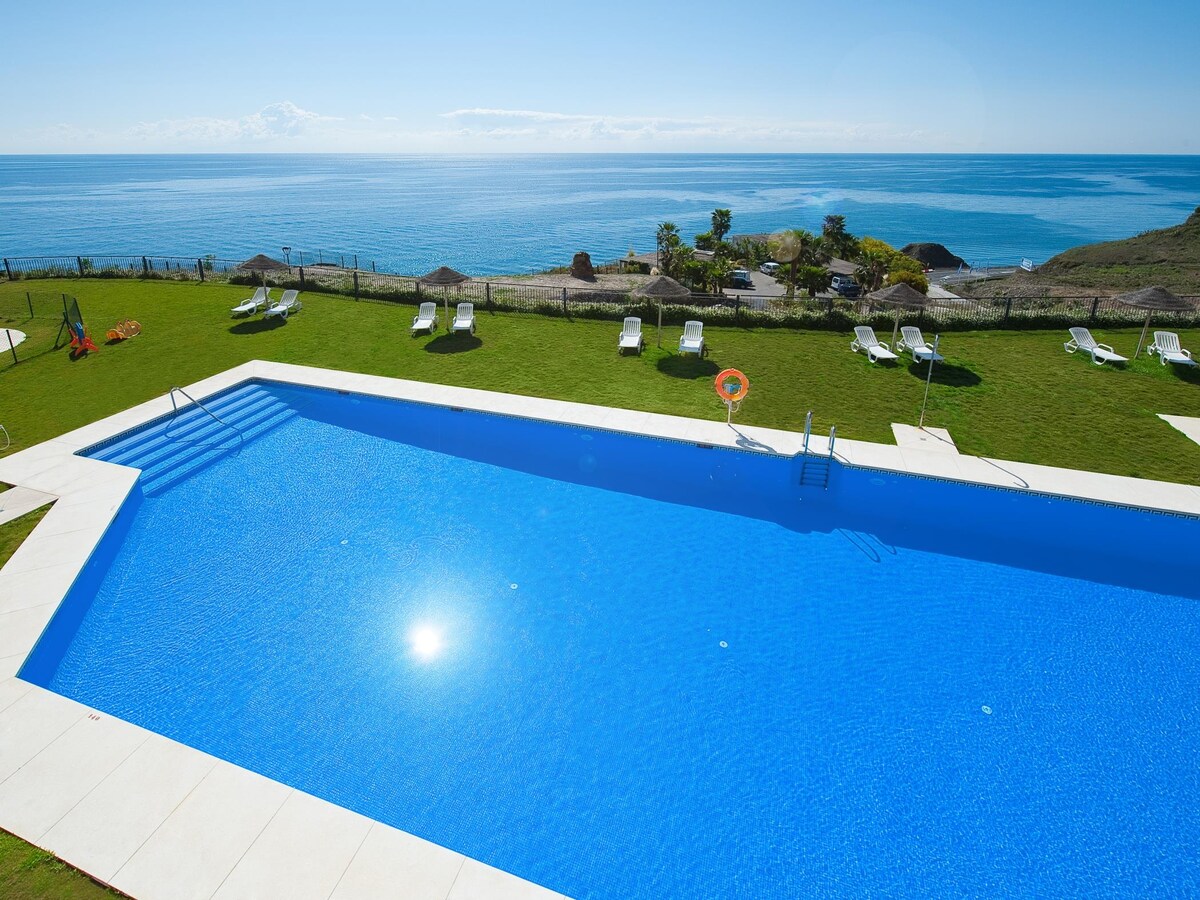 An outdoor swimming pool reflects sunlight, surrounded by well-maintained grass. Sun-loungers and sunshades are positioned nearby, providing a tranquil atmosphere. A stunning view of the ocean stretches in the background, enhancing the serene environment.