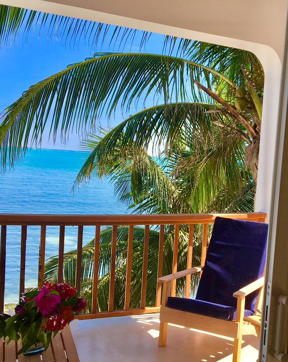A private balcony is framed by palm trees, providing a view of the sparkling ocean. A comfortable blue chair is positioned invitingly for relaxation. Vibrant flowers adorn a small table, enhancing the serene atmosphere of this coastal retreat.