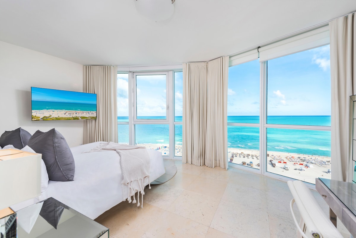 A spacious bedroom is illuminated by natural light from large floor-to-ceiling windows, offering expansive ocean views. The bed is elegantly dressed with soft linens, and a flat-screen TV is mounted on the opposite wall. Neutral-toned curtains frame the windows, enhancing the serene atmosphere.