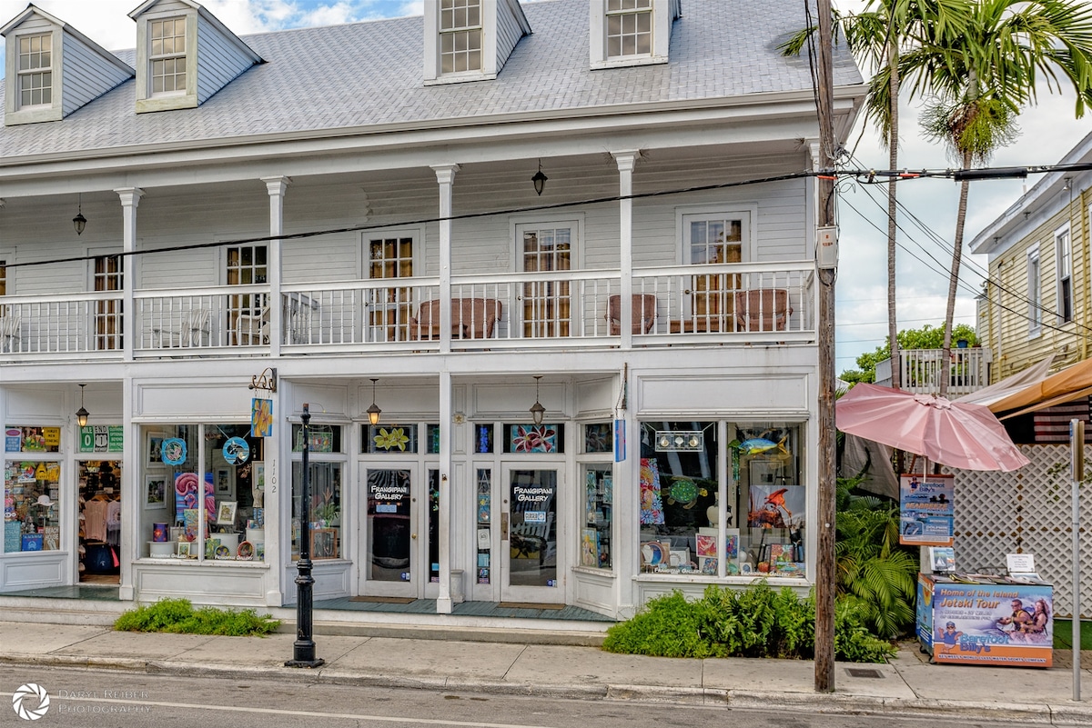 A charming white wooden building features a wraparound porch with multiple chairs overlooking the street. The ground floor showcases a variety of colorful shop windows, while the upper level includes several dormer windows under a sloped roof. Lush greenery and palm trees are visible nearby.