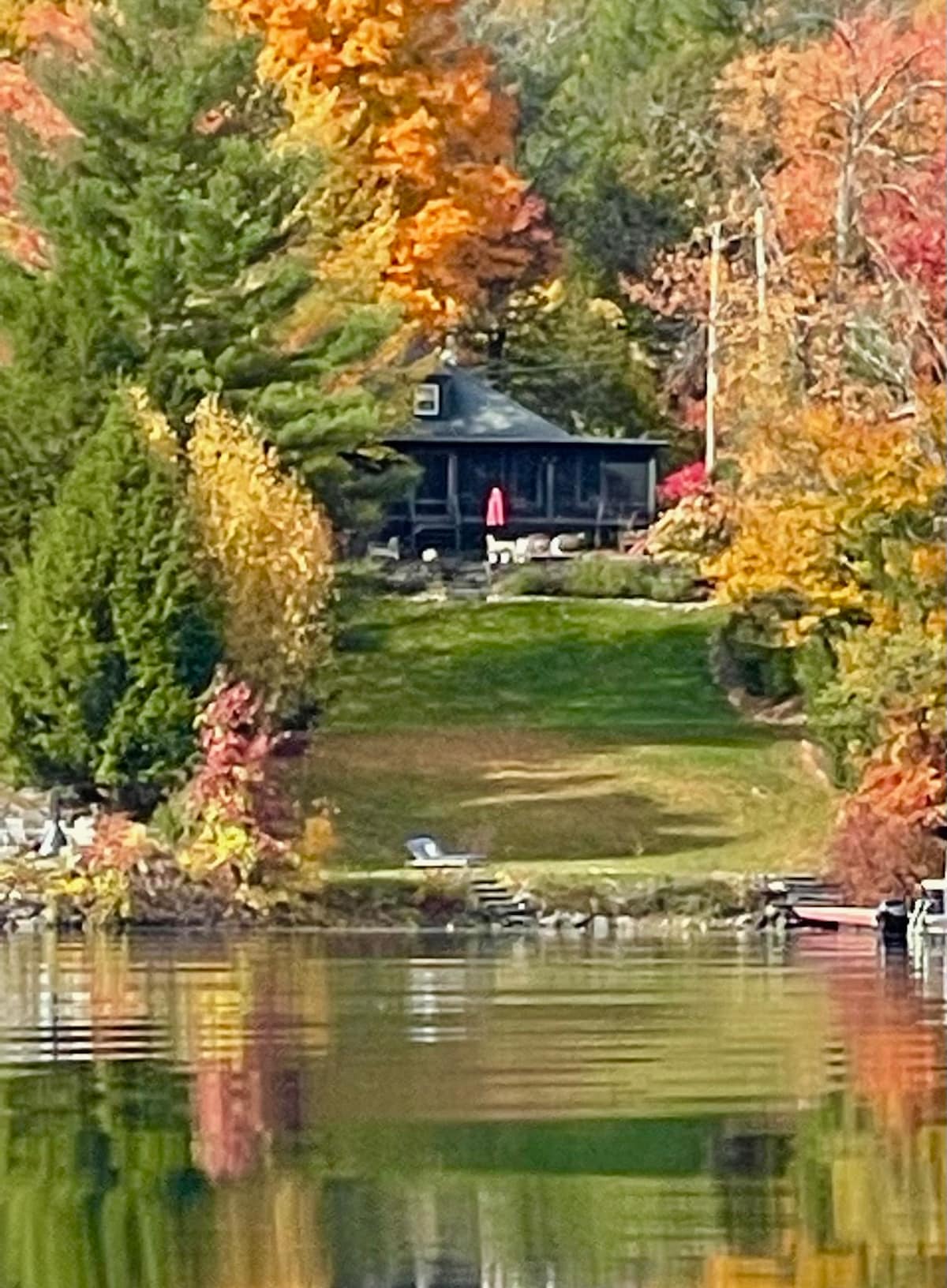 A rustic lake house is observed amidst vibrant fall foliage, with shades of red, orange, and yellow. The house features a covered porch with a bright red umbrella, while the lush green lawn slopes gently down to the water's edge, reflecting the colorful surroundings.