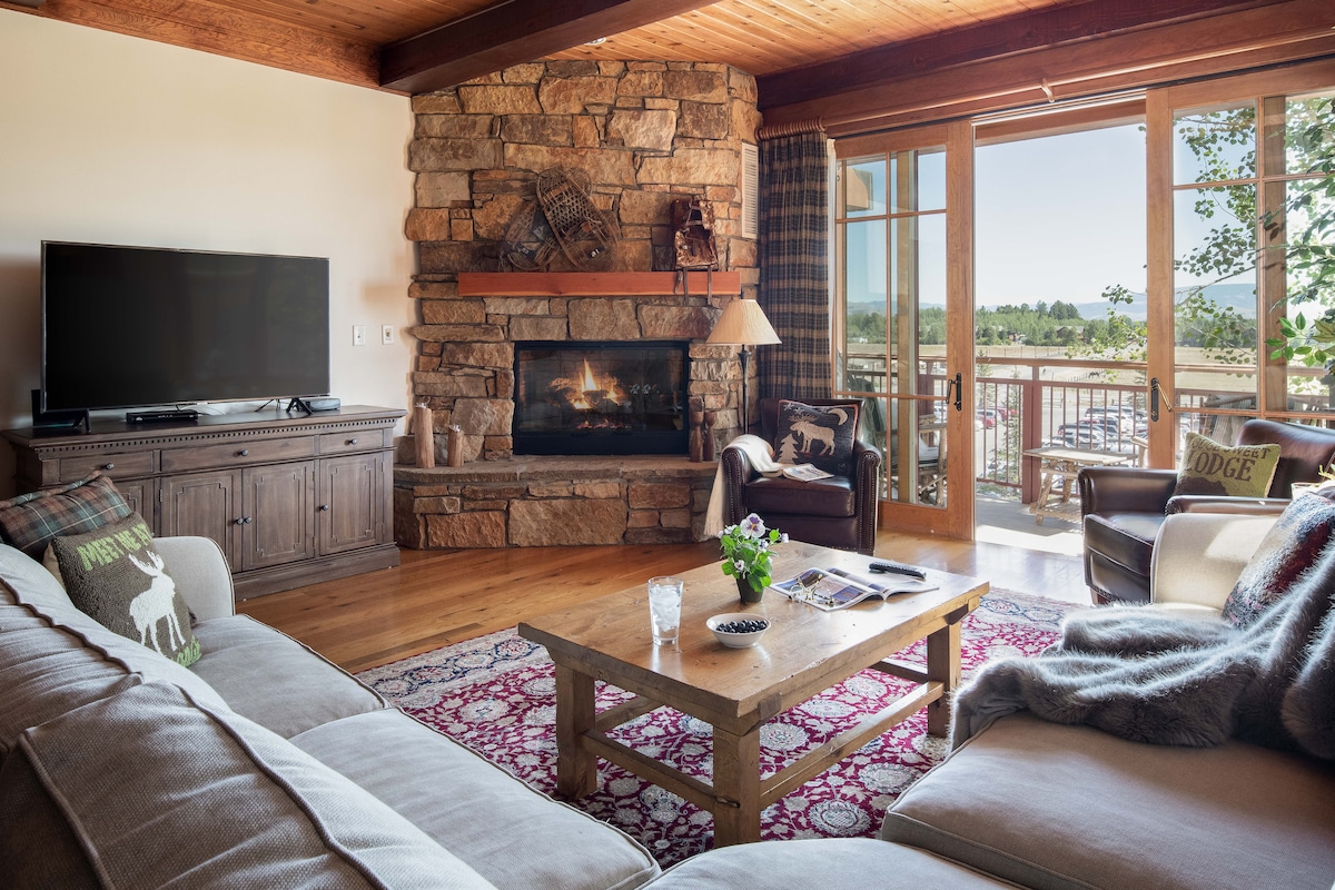 A cozy living room features a stone fireplace surrounded by comfortable seating. A wooden coffee table is placed in front of a sofa, holding magazines and a glass of water. Large glass doors lead to the balcony, offering mountain views.