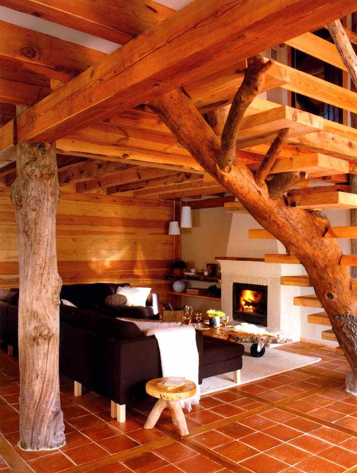 A cozy living area features wooden beams and a rustic staircase, seamlessly integrated with the natural decor. A seating arrangement with dark sofas surrounds a fireplace, providing warmth. Natural light filters through the space, enhancing the inviting atmosphere.