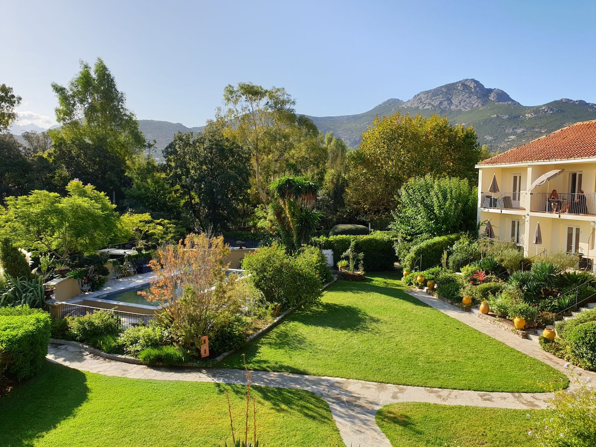A landscaped garden is viewed, featuring lush greenery, trees, and colorful flower pots. The scene includes a pathway winding through the grass, leading towards a swimming pool, with mountains visible in the background under a clear blue sky.